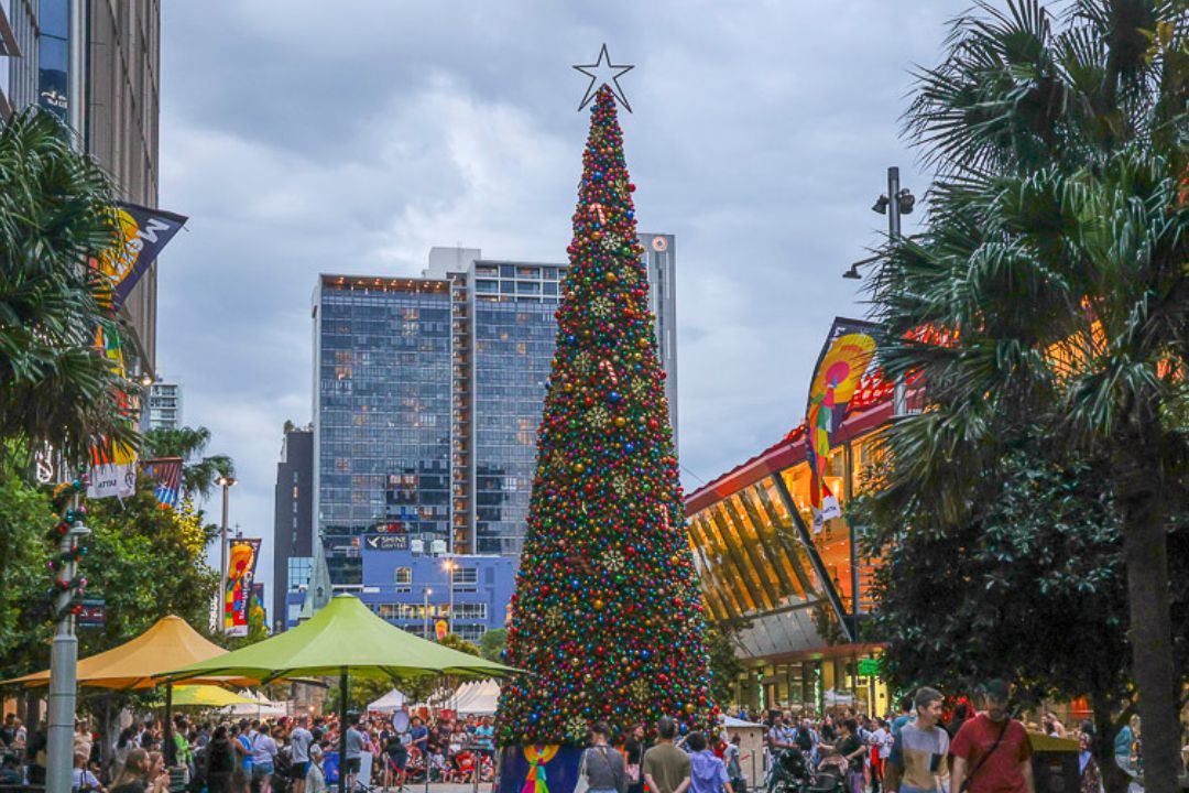 A tall Christmas tree adorned with lights stands in a lively city square filled with people. Bright decorations and buildings create a festive atmosphere.