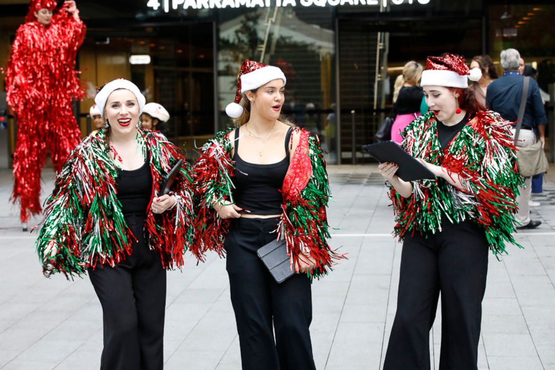 Three people in colourful fringed costumes and hats stand smiling on a street. They exude joy and creativity, with a festive and lively atmosphere.