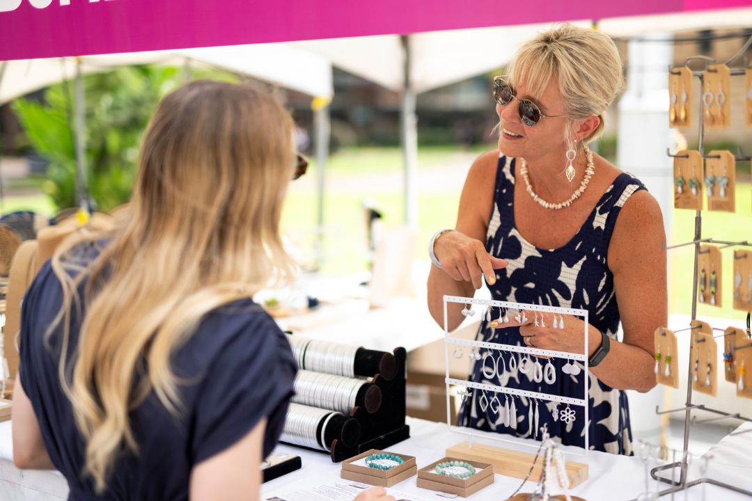 Two women in a lively outdoor market setting, one buying jewellery from the other. The seller smiles warmly, showcasing a necklace under a pink canopy.
