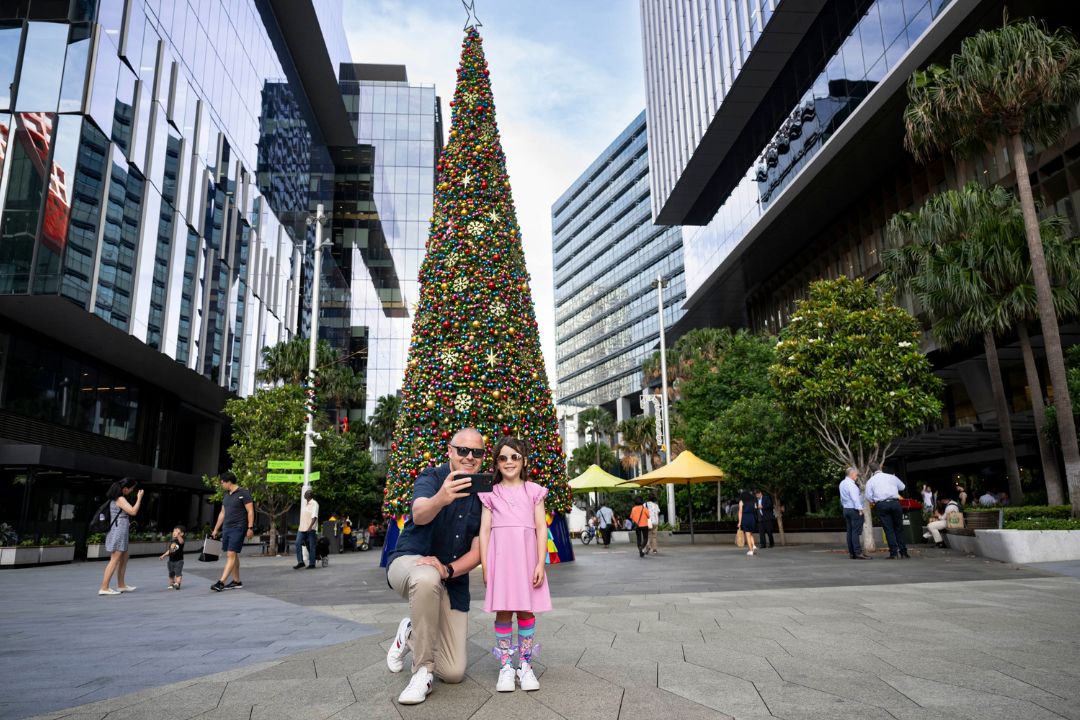 A towering Christmas tree adorned with ornaments stands in a bustling urban plaza, surrounded by skyscrapers. People walk and take photos, conveying holiday cheer.