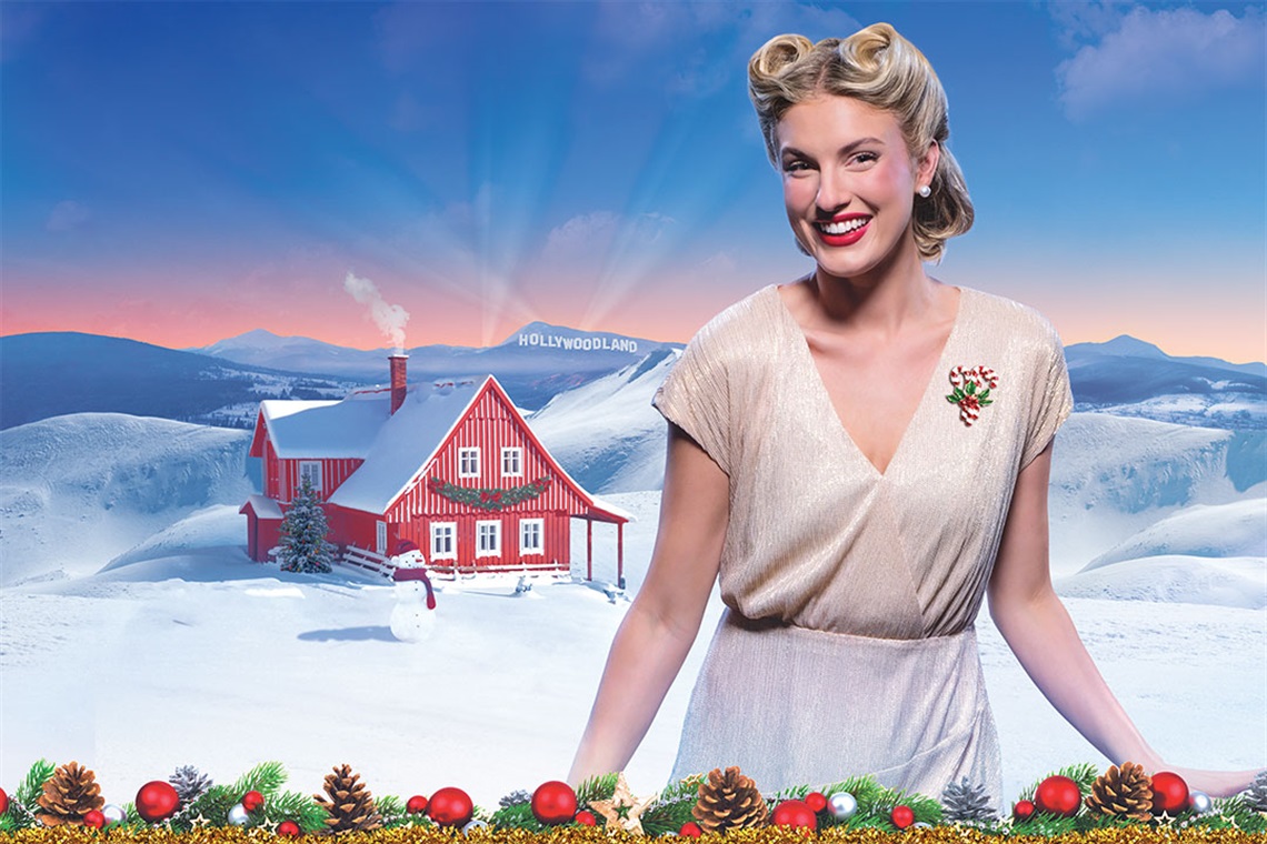 A smiling woman in vintage clothing stands in a snowy landscape with a quaint red-roofed house. The scene conveys a cheerful, nostalgic winter feel.