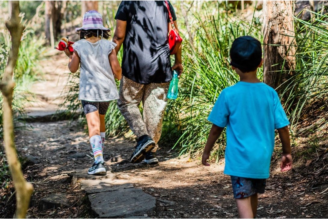 Two children walk along a sunny forest trail, accompanied by an adult. The scene conveys a sense of exploration and adventure in a natural setting.