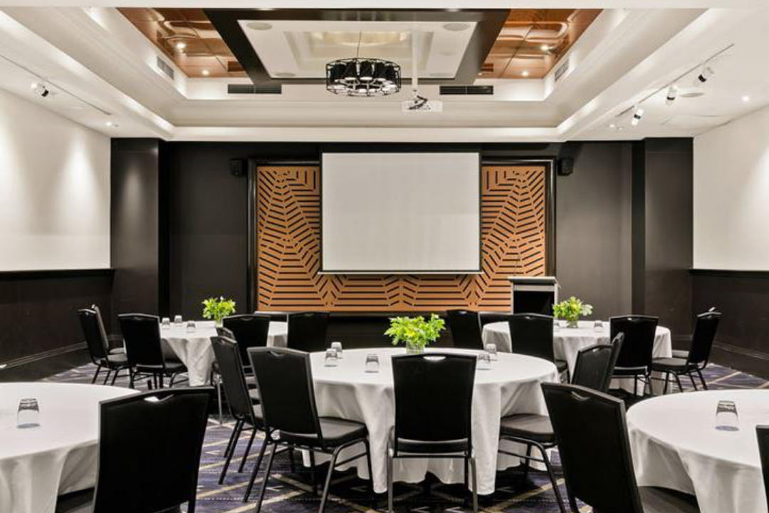 Image of conference facilities within a hotel in Parramatta. Pictured are round tables and chairs with a lectern and presentation screen