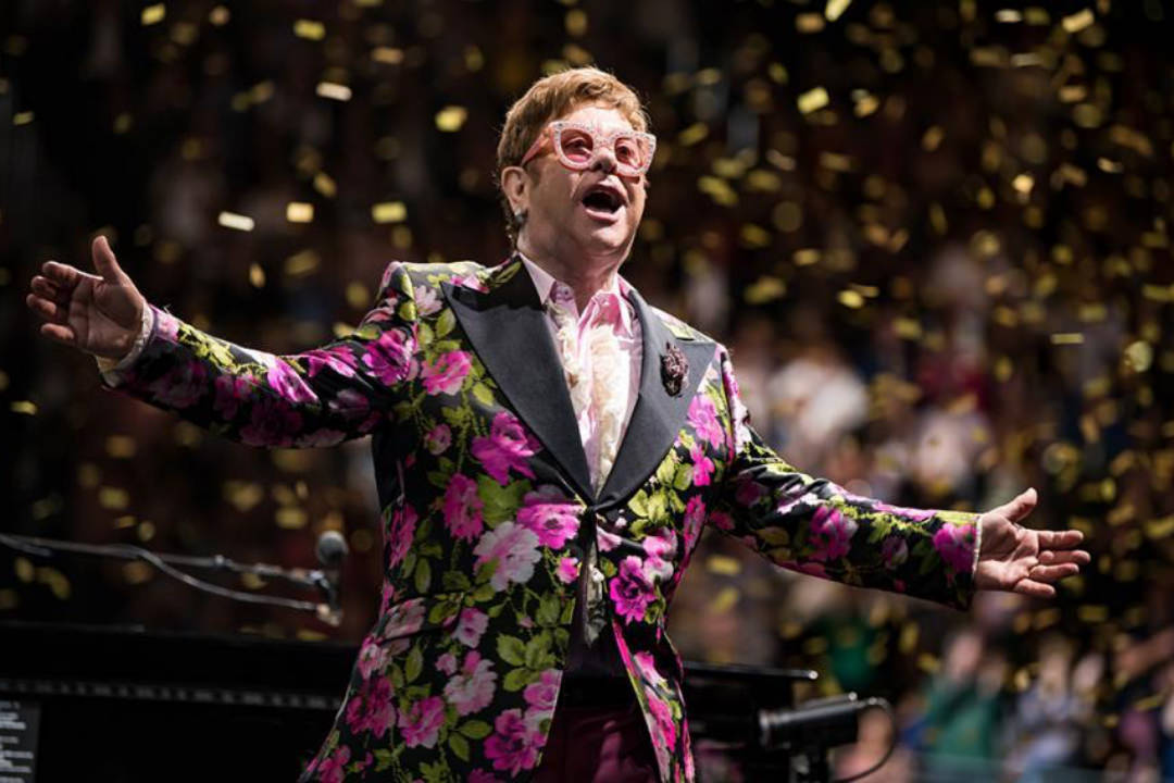 Image of Sir Elton John dressed in a flamboyant green pink and black blazer standing in front of his piano with his arms open about to take a bow. 