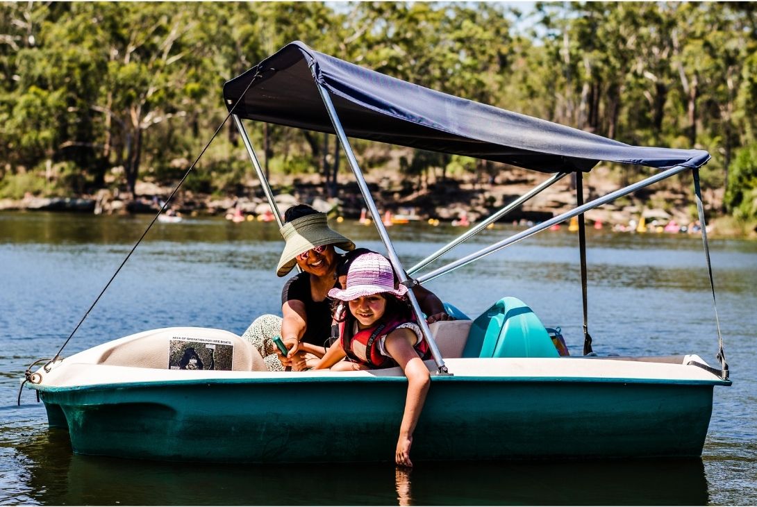 Two people in hats and sunglasses relax on a small boat with a canopy, drifting on a calm lake surrounded by trees. A peaceful and sunny day.
