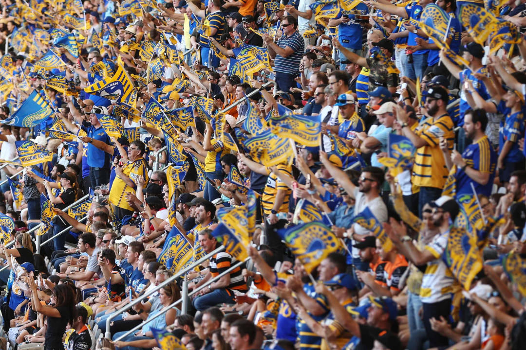 A Parramatta Eels player making a play at the ball while an opposing team player goes in for a tackle. In the background, blurred out, are Parramatta Eels fans. 