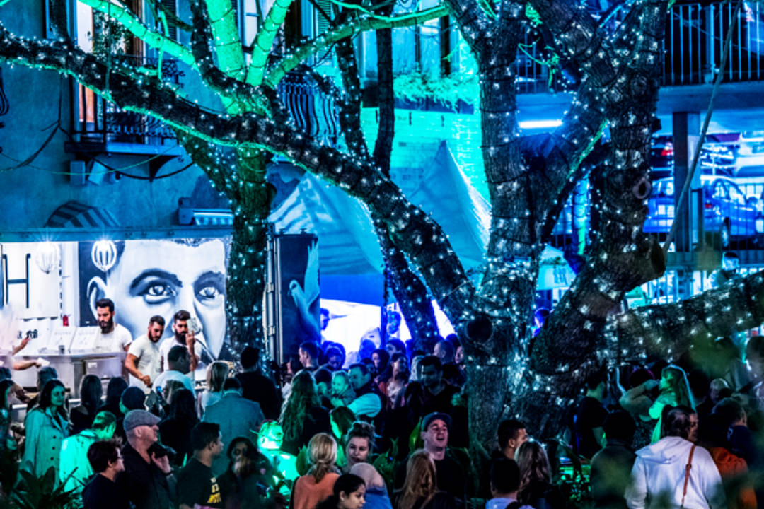 Revelers and partying people dance and socialise around a DJ in an activated space on Phillip Street. The trees and buildings are glowing from a blue-green floodlight effect and there are murals of famous DJs project onto buildings. 