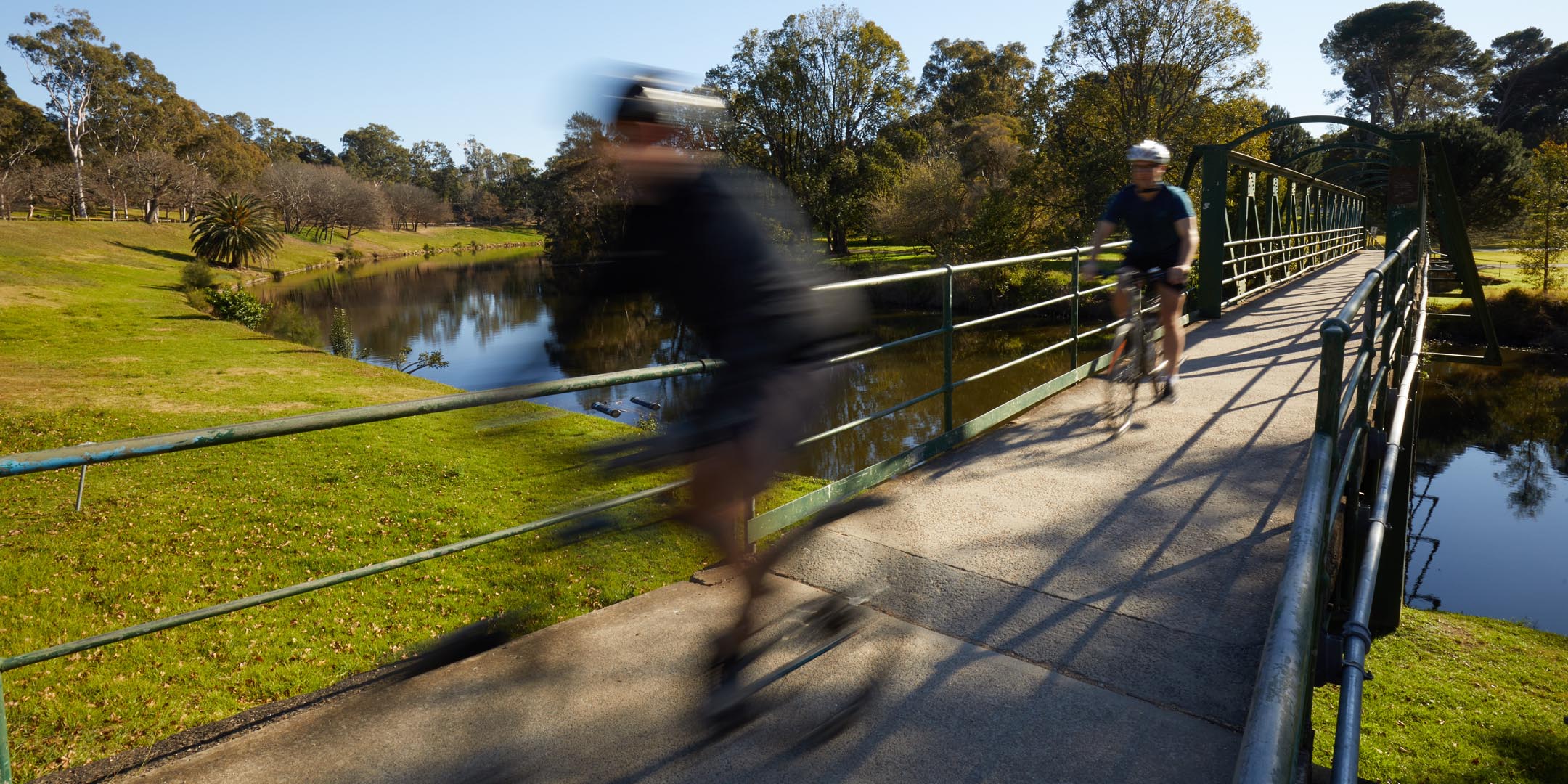 two cyclists are shown crossing a bridge over the Parramatta River into Parramatta Park. To illustrate the effect of speed the 1st cyclist has been blurred. 
