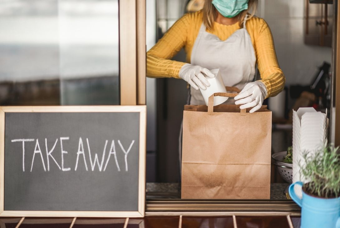 cafe worker wearing mask and gloves prepping takeaway bag with a takeaway sign next to her