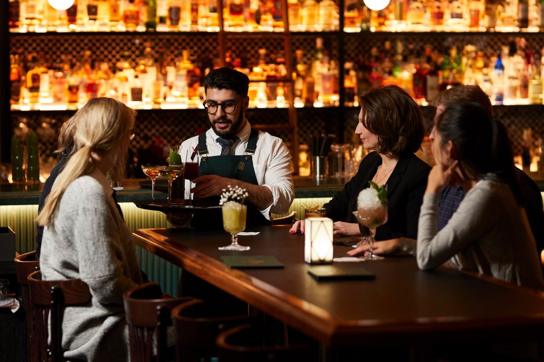 four people sitting around a table in a bar while a waiter hands out cocktails