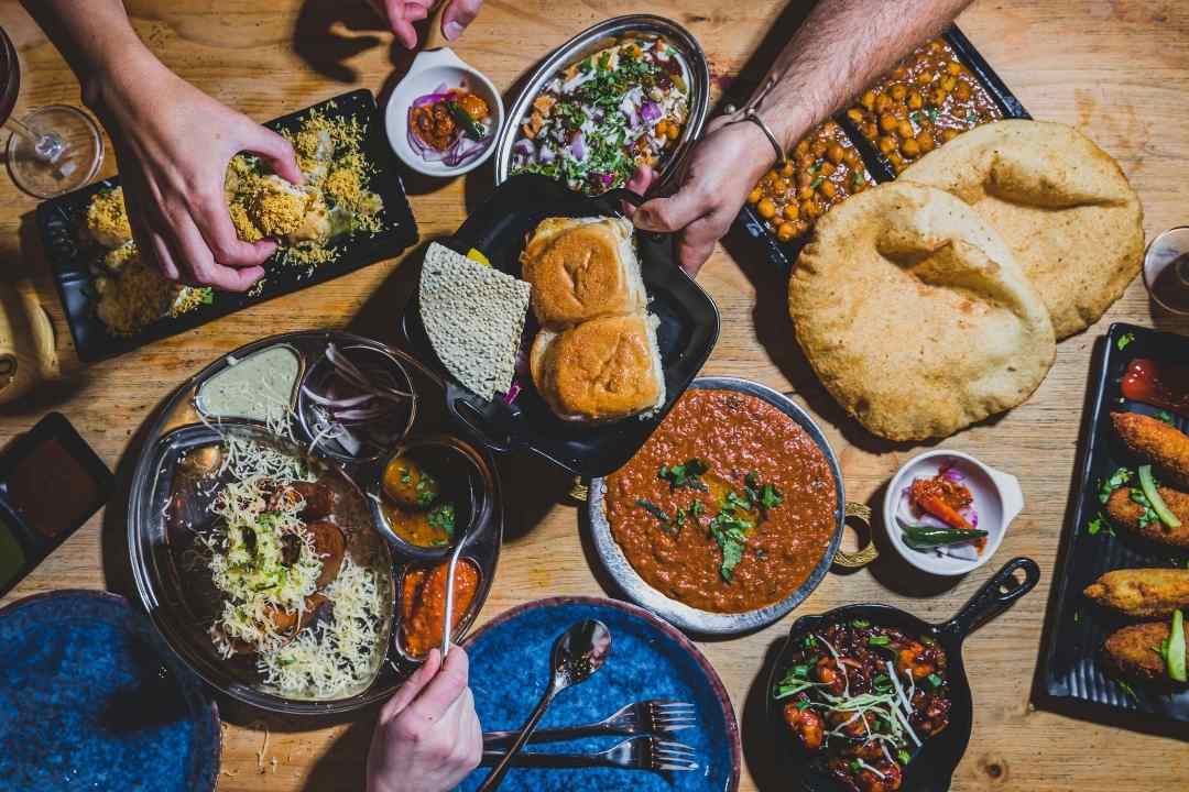 Top view of a table filled with various Indian dishes, including curries, flatbreads, dips, and garnished plates. Hands are reaching for food, evoking a communal and vibrant atmosphere.