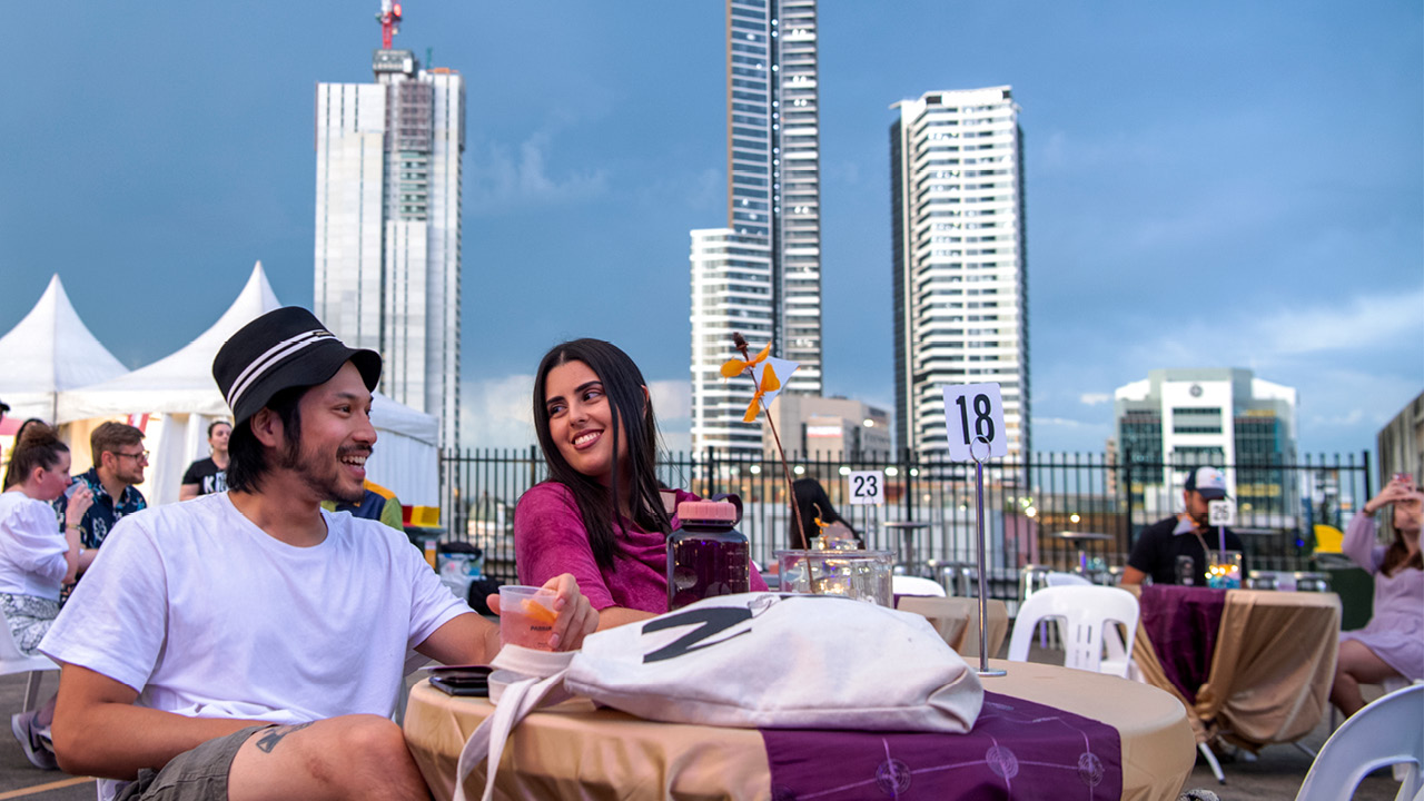 Two people sitting at a table on a roof top with tall buildings behind them.