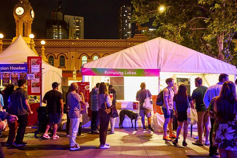 Night market scene with people gathered outside a lit food stall labeled "Kampung Laksa." Festive atmosphere with city buildings and a clock tower.
