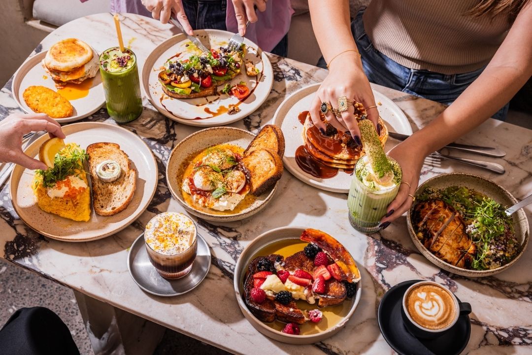 Aerial view of a table filled with diverse dishes, including burgers, pancakes, and salads. People are reaching in, creating a lively, shared meal atmosphere.