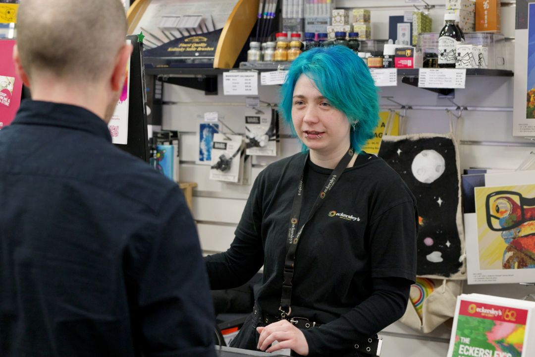 Woman in blue hair and wearing black shirt standing in front of counter with art supplies behind her like paint tubes