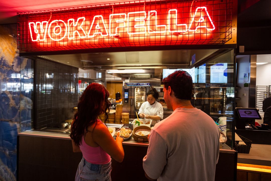 Man and woman standing at counter under the sign "Wokafella"