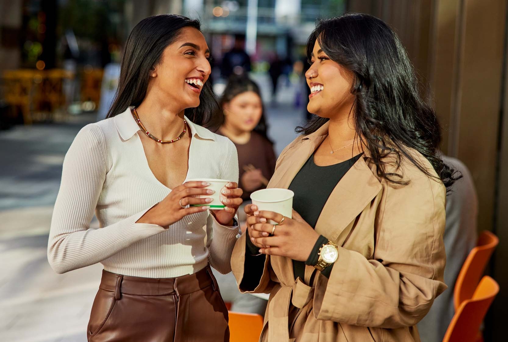 Two friends laugh and chat together in Parramatta Square while holding takeaway cups of miso soup.