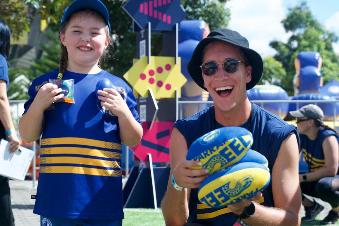 A child and an adult, both smiling and wearing blue sports attire, pose outdoors with rugby balls. The scene is lively and festive with bright colors.