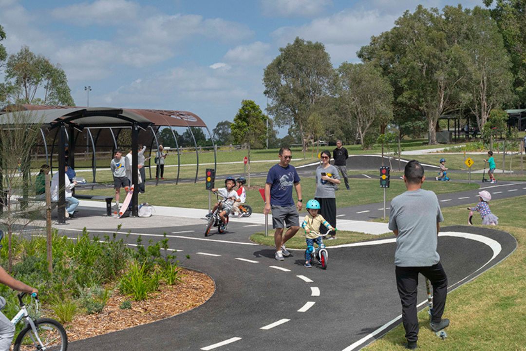 Children ride bikes on a winding path in a park. Adults watch nearby. Trees and a clear sky provide a serene backdrop, adding to the cheerful scene.