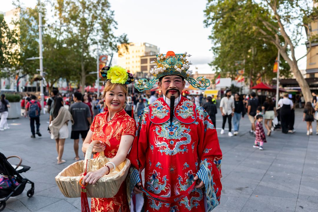 A couple dressed as bright red lobsters holds baskets, smiling on a lively street with people and greenery around. The atmosphere is festive and cheerful.