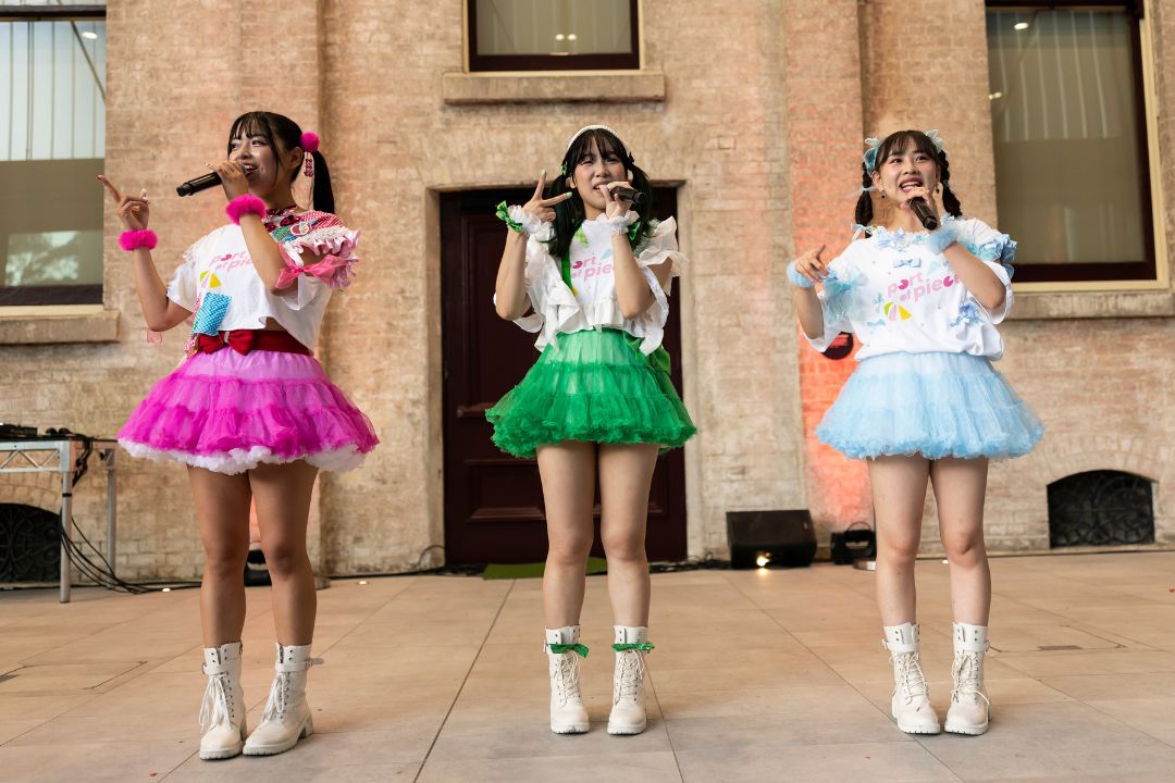 Three women perform outdoors, wearing colorful skirts in pink, green, and blue with white tops. They sing into microphones, smiling energetically.