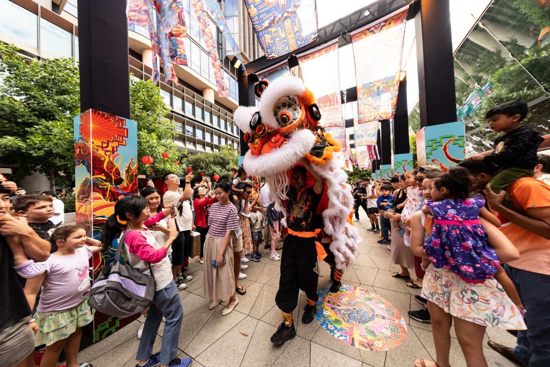 A lively street performer dressed in colorful costume entertains an excited crowd. Children and adults watch eagerly, creating a festive atmosphere.