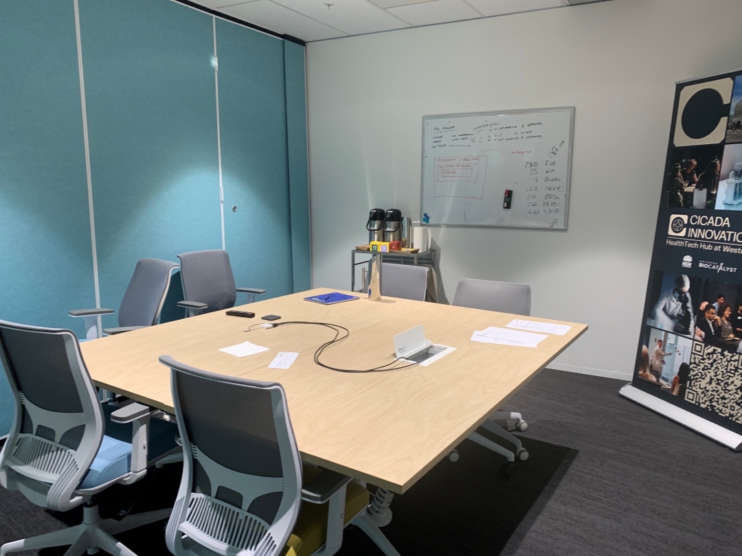 A modern conference room with a wooden table and six office chairs. A coffee machine sits on a side cabinet. The room feels organized and professional.