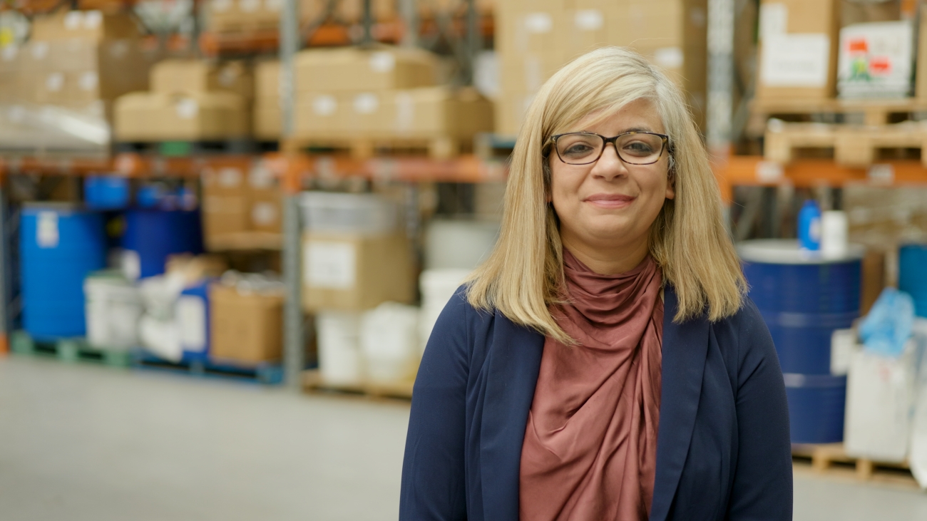 A woman with long blonde hair and glasses stands in a warehouse, surrounded by stacked boxes and supplies. She looks content and professional.