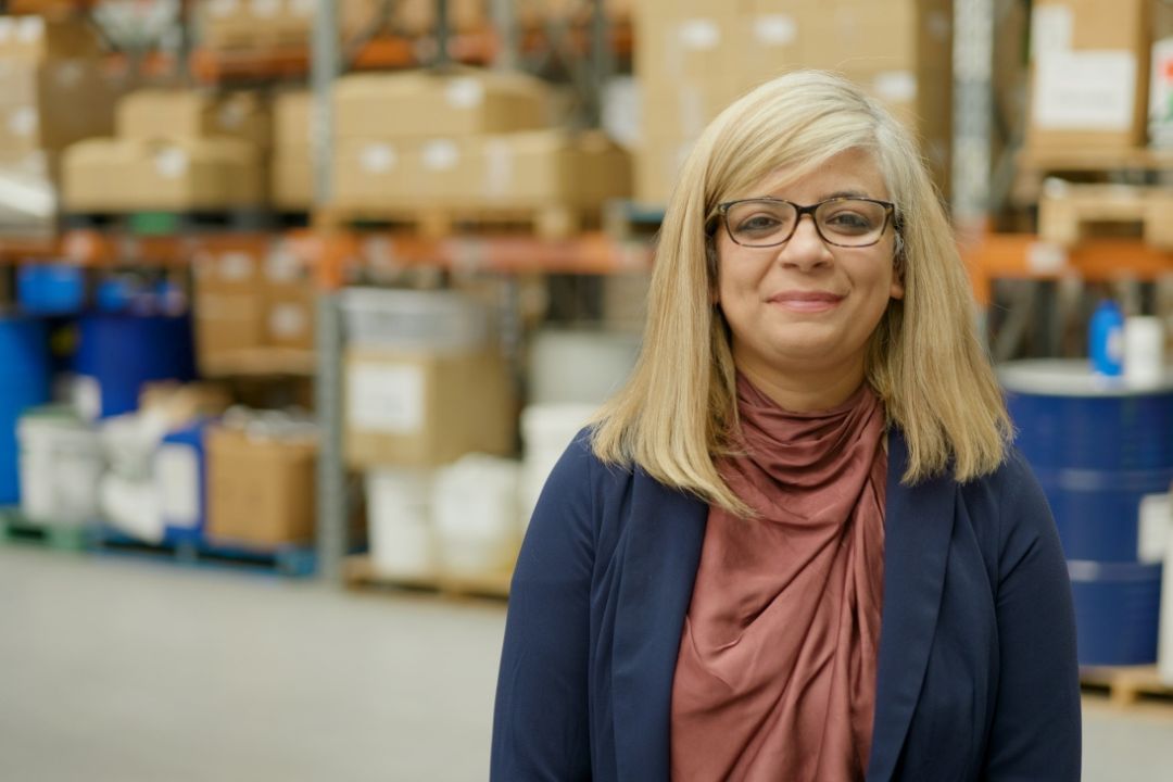 A woman with long blonde hair and glasses stands in a warehouse, surrounded by stacked boxes and supplies. She looks content and professional.