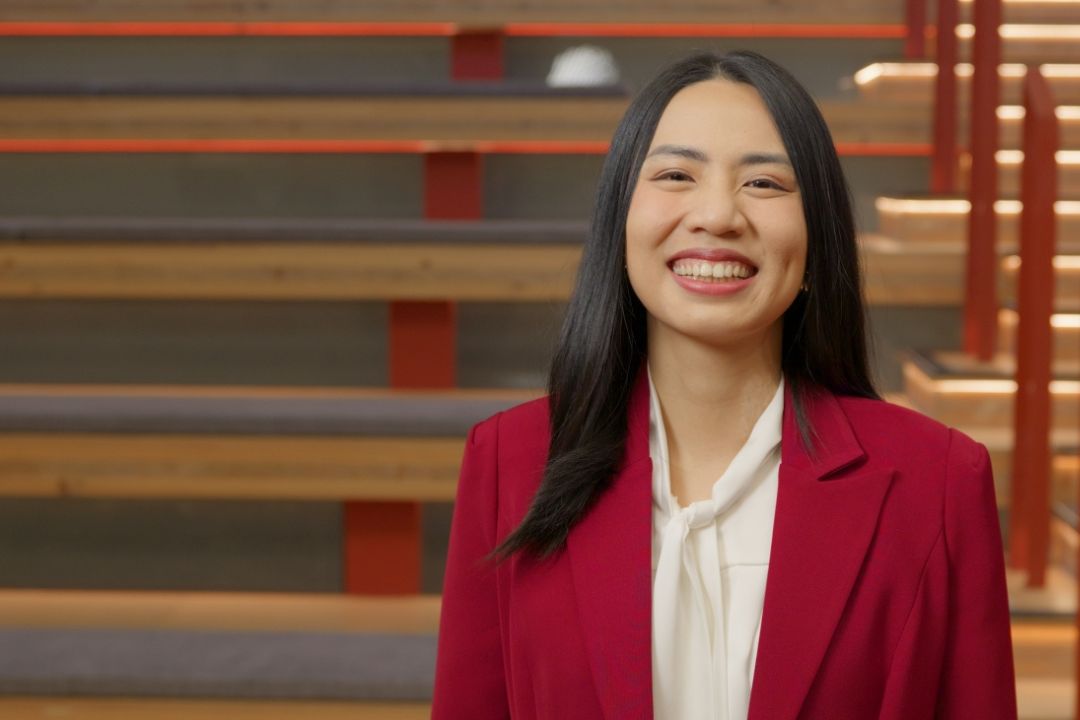 A woman in a red blazer and white blouse smiles warmly. She stands in front of wooden blinds, creating a professional and welcoming atmosphere.