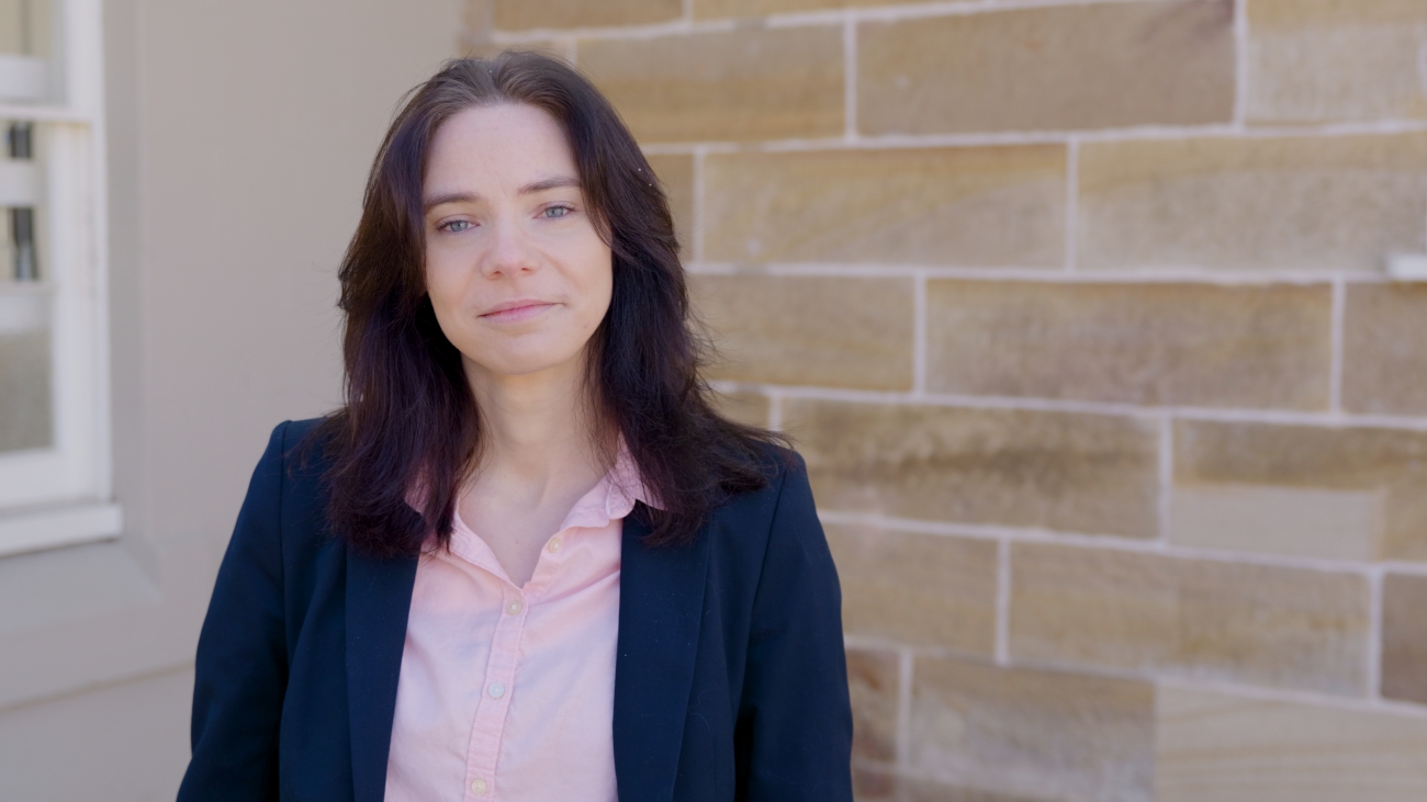 A woman with dark hair wearing a black blazer and pink top stands in front of a beige brick wall, looking confidently at the camera. Bright, natural lighting.