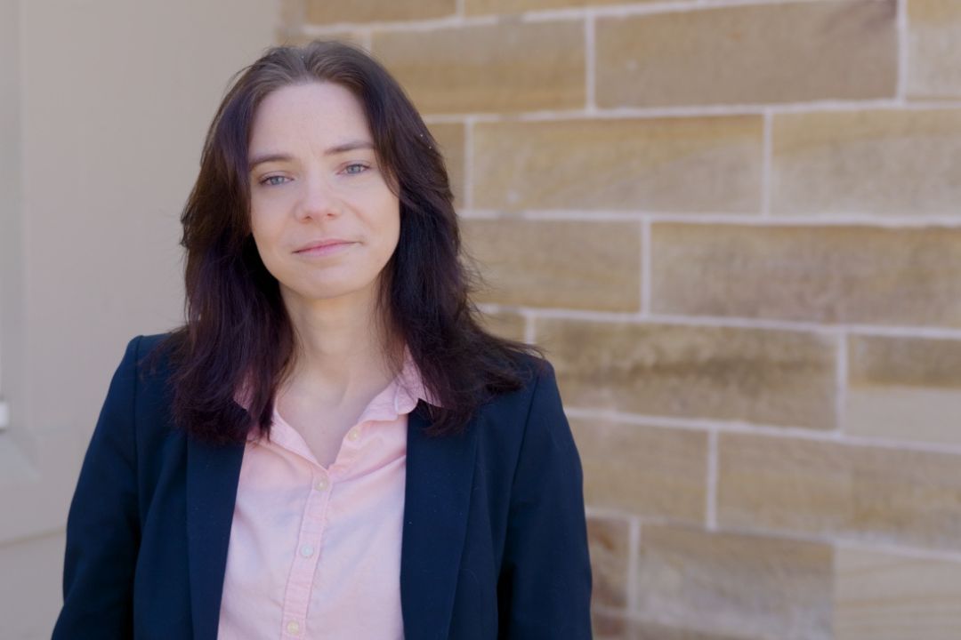 A woman with dark hair wearing a black blazer and pink top stands in front of a beige brick wall, looking confidently at the camera. Bright, natural lighting.