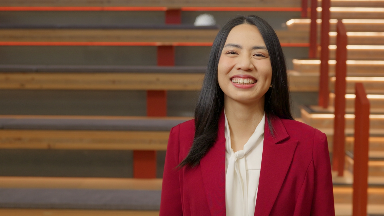 A woman in a red blazer and white blouse smiles warmly. She stands in front of wooden blinds, creating a professional and welcoming atmosphere.