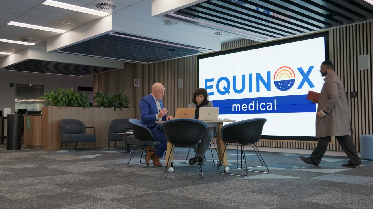 Modern office space with people interacting. A man in a suit sits at a table with a laptop. A large screen displays the "Equinox Group" logo.