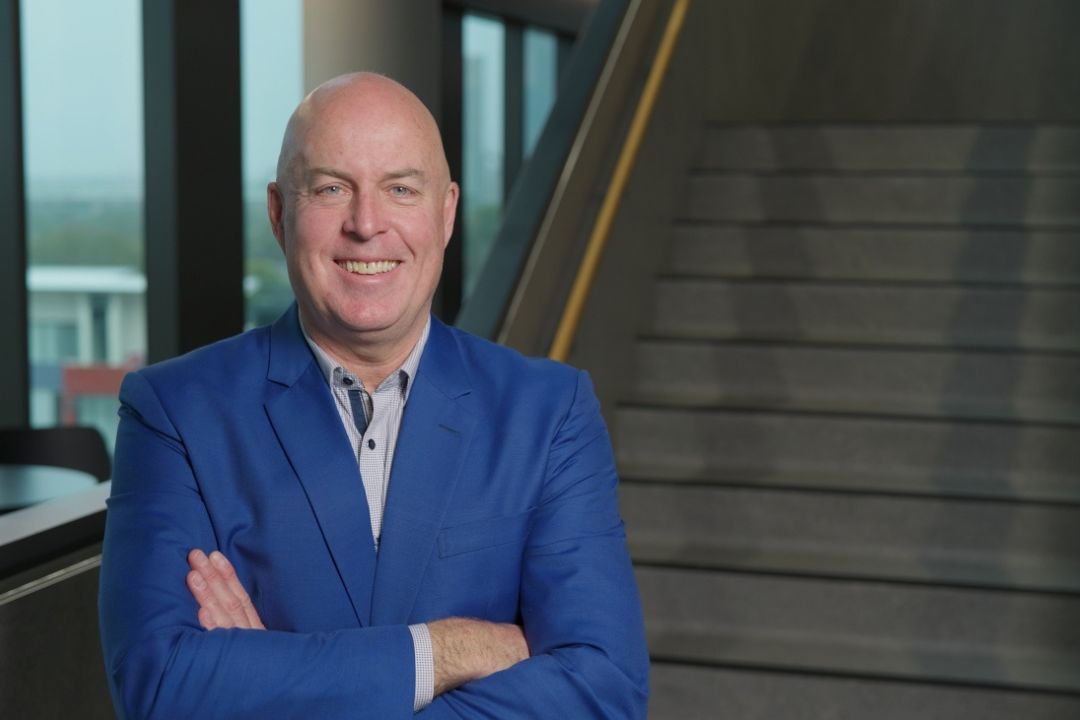 A man in a blue blazer stands confidently with crossed arms in a modern, well-lit building. Behind him, a wide staircase ascends, creating a professional tone.