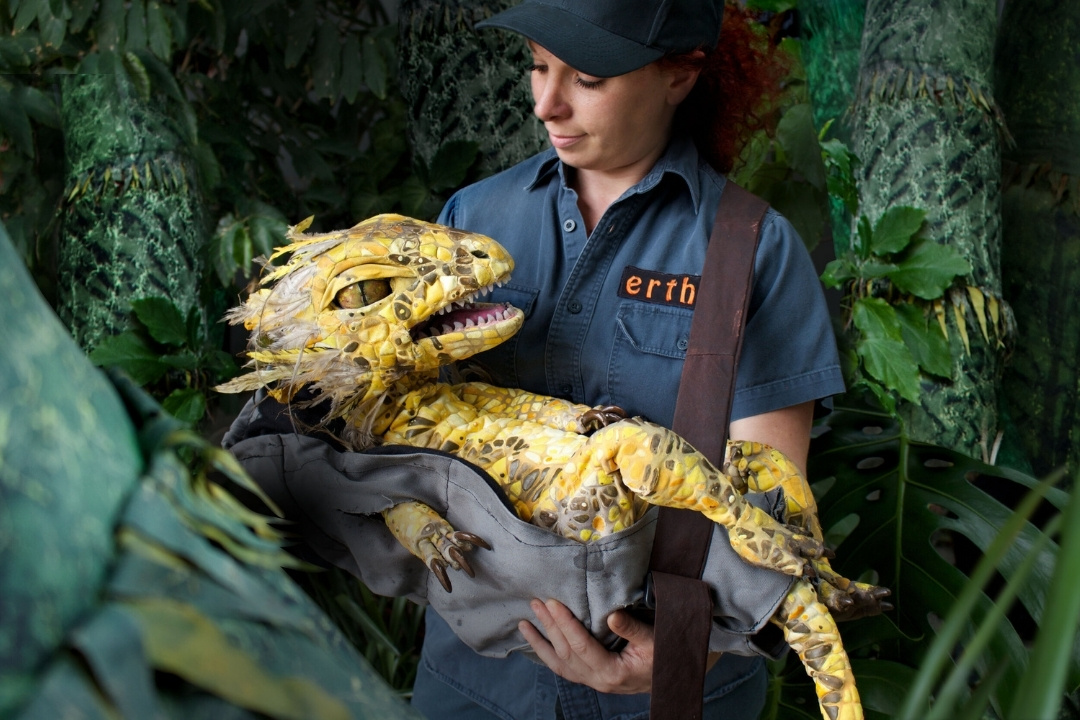 A person in a blue outfit holds a lifelike yellow dinosaur puppet in a lush jungle setting. The atmosphere is adventurous and imaginative.