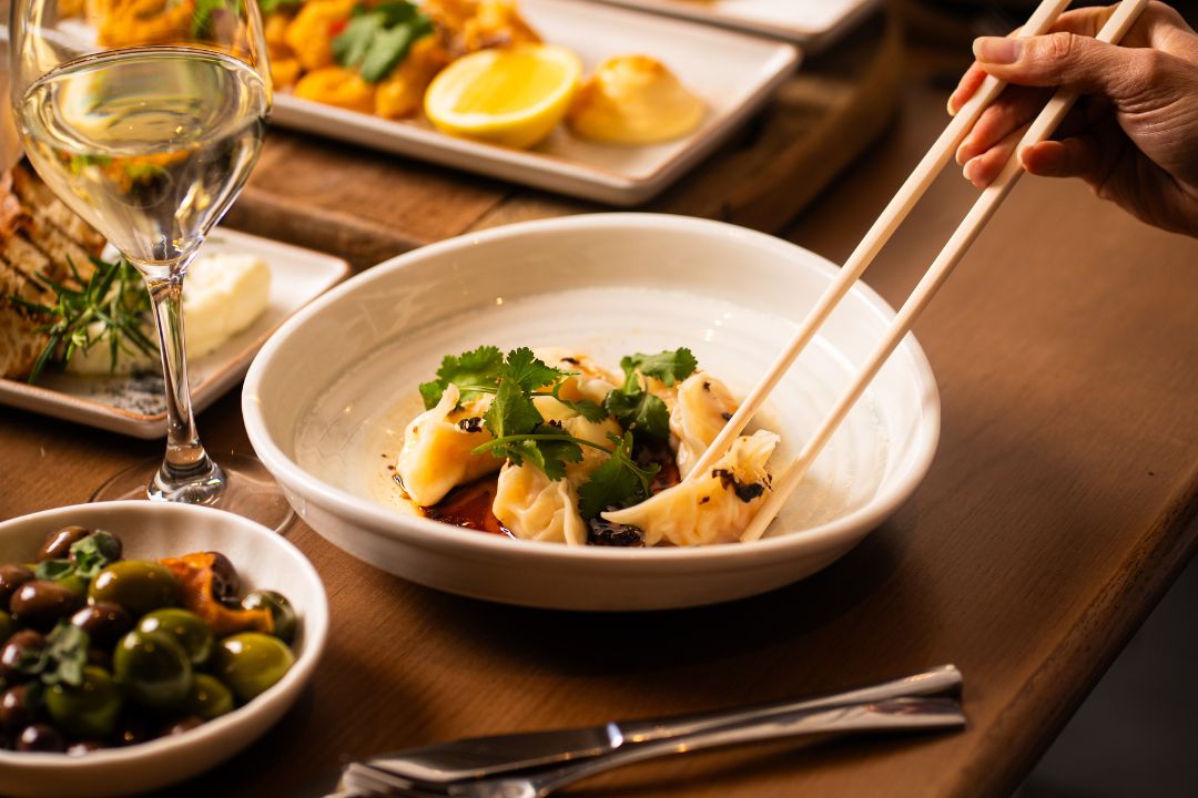A hand uses chopsticks to pick up a dumpling from a white bowl garnished with cilantro and sauce.