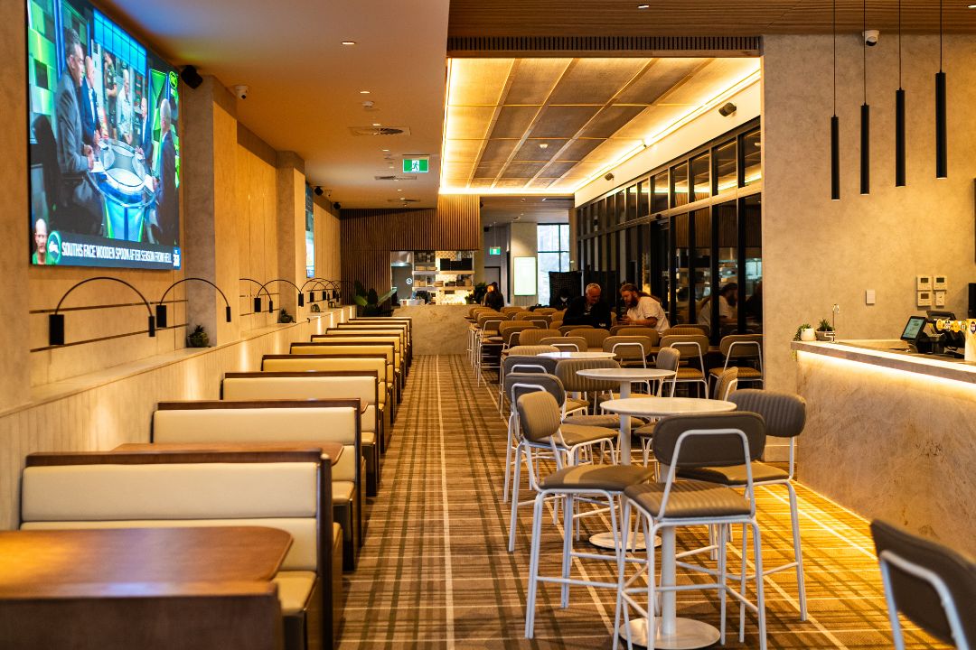 Modern restaurant interior with booth seating on the left, round tables and chairs in the center. A large TV screen displays a talk show.