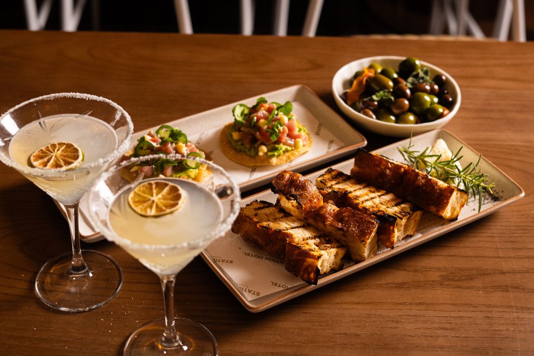 Two martinis with dried lime, grilled bread with herbs, a vibrant tostada with avocado, and a bowl of assorted olives on a wooden table.