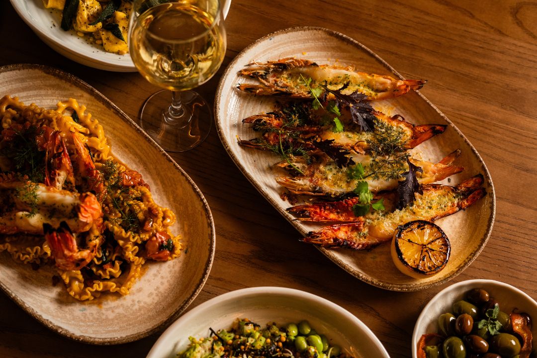 A table setting features plates of grilled prawns and pasta with shrimp, a glass of white wine, and bowls of olives and edamame