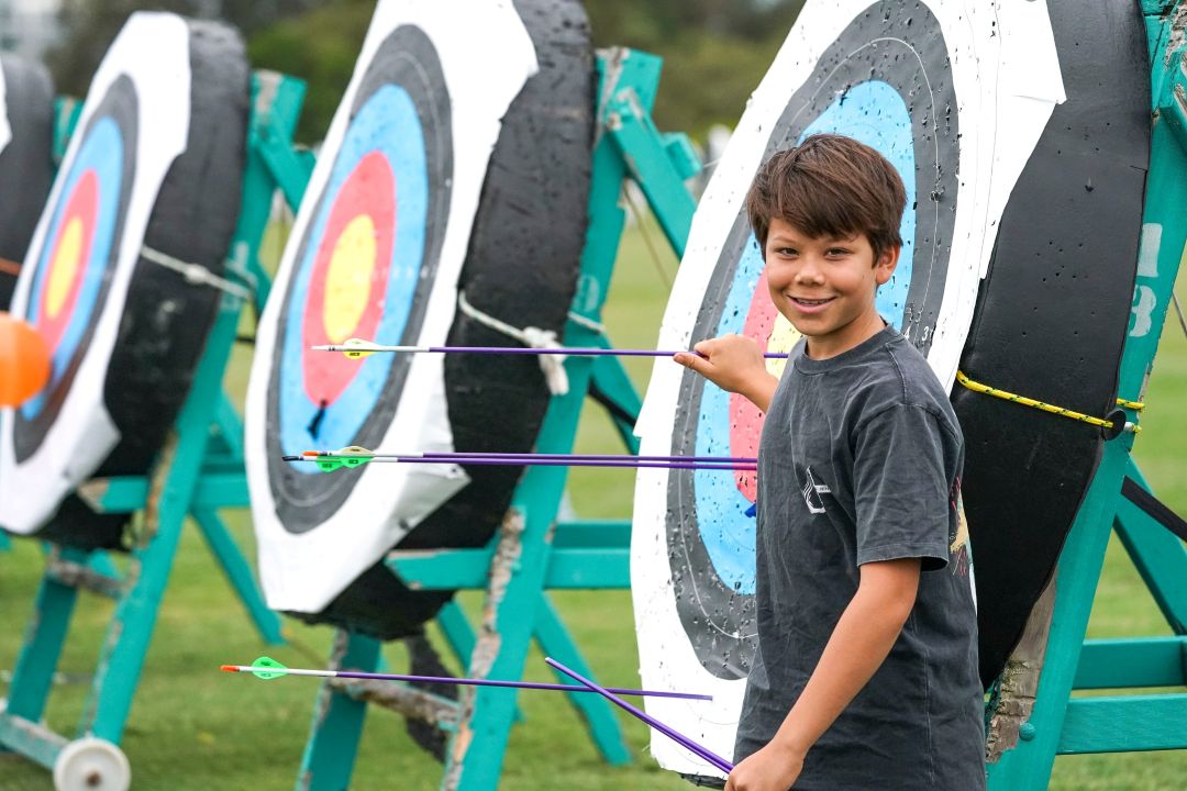Smiling boy stands near archery targets with arrows embedded in the bullseyes, outdoors on a grassy field. The scene conveys achievement and joy.