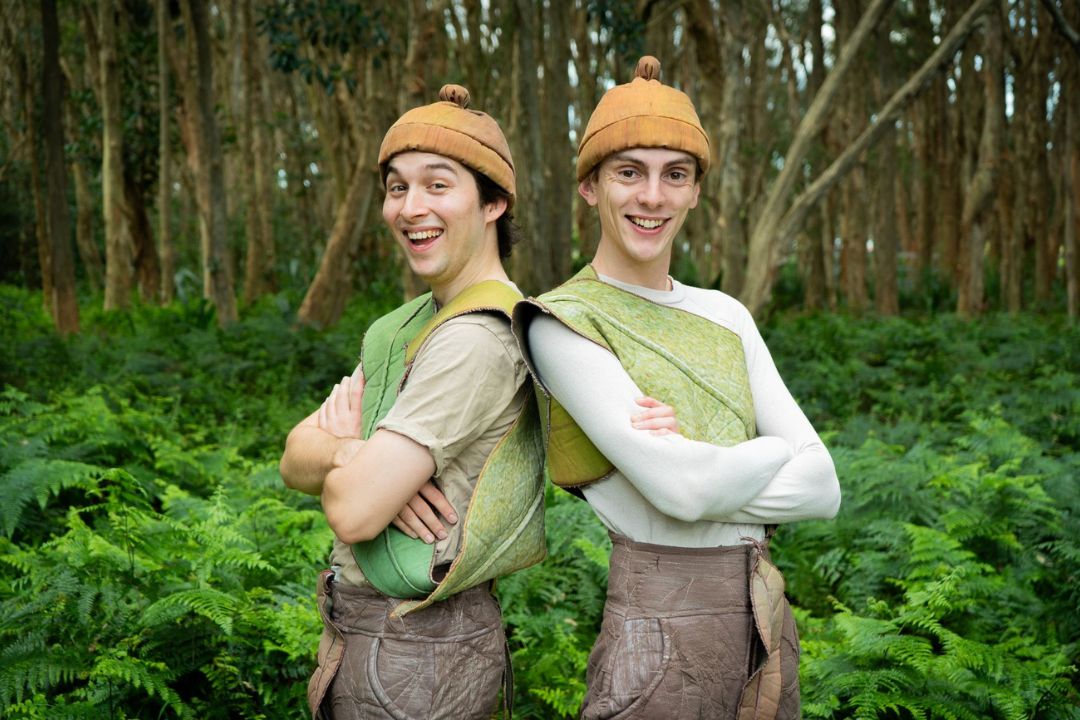 Two people in medieval-style costumes stand back-to-back in a lush green forest, smiling confidently. They wear brown hats and green tunics, blending with the surroundings.