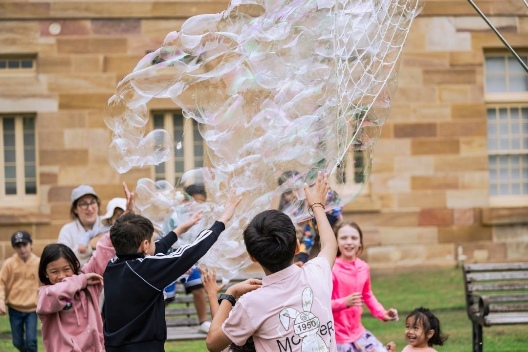 Children joyfully chase large bubbles on a green lawn in front of a stone building. Their expressions are playful and excited, capturing a moment of carefree fun.