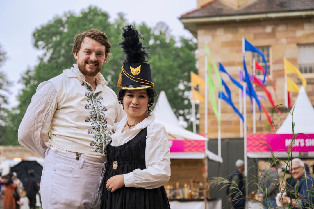 Two people in historical costumes pose cheerfully outside a building with flags. The scene conveys a festive and celebratory atmosphere.