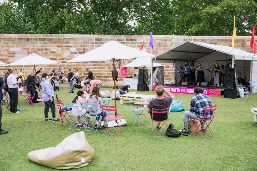 Outdoor music festival with people sitting on the grass, enjoying a live band on stage. White tents and trees create a relaxed, festive atmosphere.