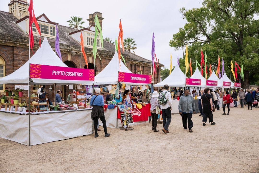 Outdoor festival scene with people walking past food stalls under white tents, adorned with pink banners. The atmosphere is lively and relaxed.