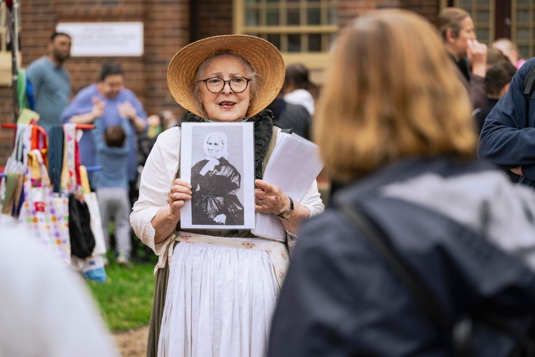 A woman in period costume and straw hat joyfully displays a vintage photo to an engaged crowd outdoors, suggesting a historical reenactment event.