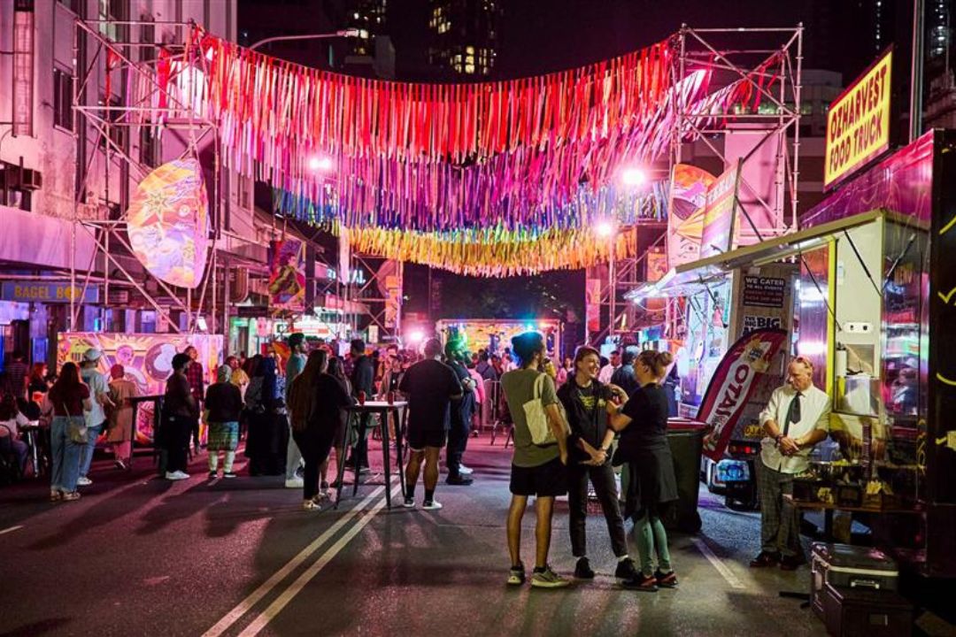 A vibrant street scene at night, with colorful hanging decorations and pink lighting. People walk and gather near food stalls, creating a lively atmosphere.