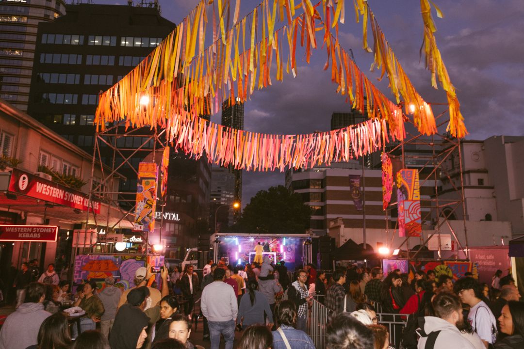 A lively street festival at dusk, with colorful lights and decorations overhead. A crowd gathers, enjoying a festive atmosphere in an urban setting.