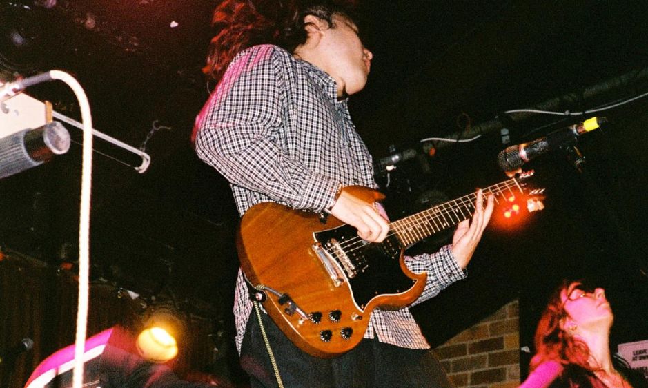 A musician energetically plays a guitar on stage, surrounded by colorful stage lights. The atmosphere is lively and dynamic, capturing the essence of a live performance.